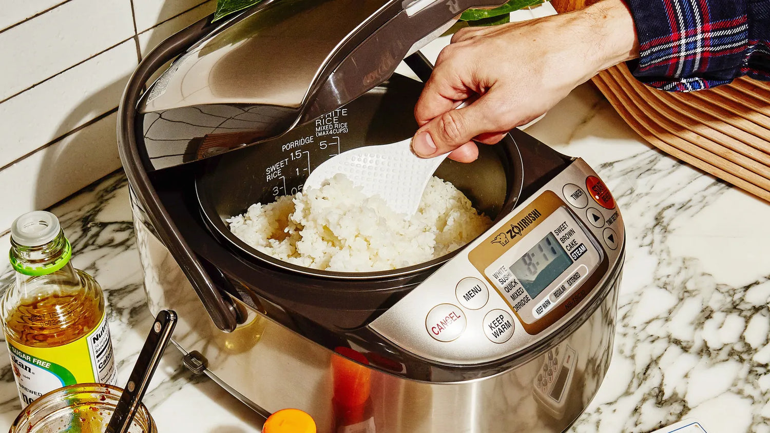 A hand is scooping out cooked white rice from a rice cooker with a paddle.